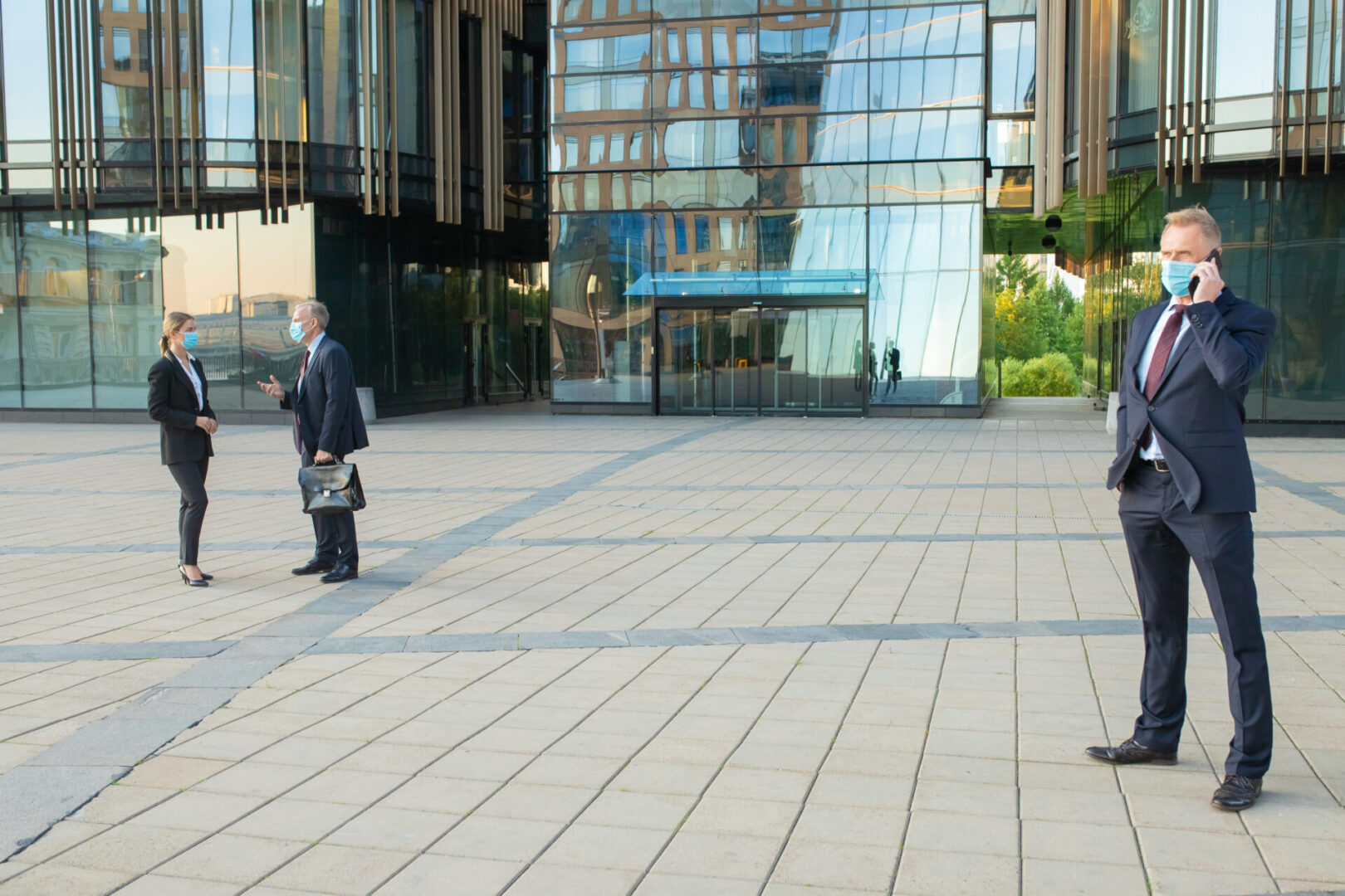 Businesspeople wearing masks and suits, talking at office building, speaking on cellphone, discussing project with each other. Copy space, full length. Business and epidemic concept