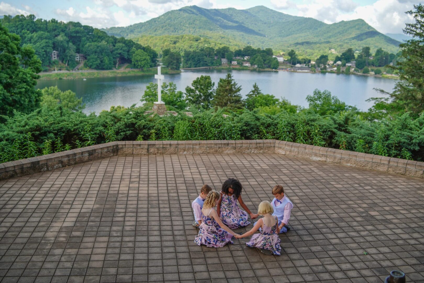 A group of kids praying near a cross surrounded by a lake and hills covered in forests